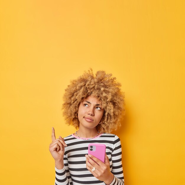La giovane donna europea dai capelli ricci pensierosa punta sopra su uno spazio vuoto dimostra il posto per la tua pubblicità tiene il telefono cellulare vestito con un maglione a righe casual isolato su sfondo giallo