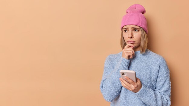 La giovane donna europea frustrata con espressione stressata tiene il mento sembra sconvolto usa le chat del telefono cellulare online ha problemi isolati su sfondo marrone spazio di copia per la tua promozione.