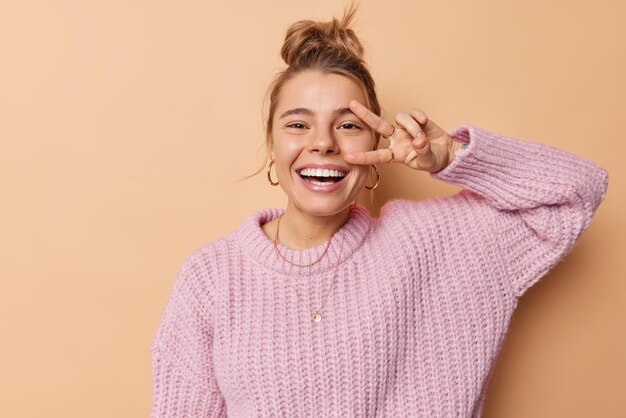 La giovane donna europea positiva fa segno di pace sulle risate degli occhi con gioia si gode la vita indossa un maglione lavorato a maglia sciolto isolato su sfondo marrone dello studio. Gesto di vittoria. Concetto di linguaggio del corpo