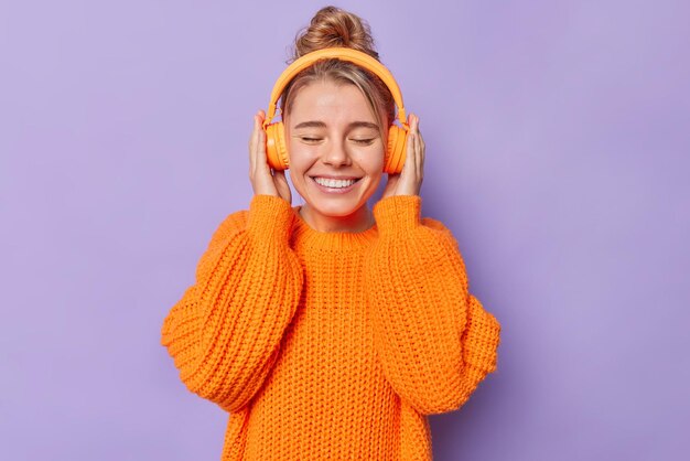 La giovane donna europea positiva tiene le mani sulle cuffie stereo ascolta la traccia audio sorride a denti stretti indossa un caldo maglione lavorato a maglia isolato su sfondo viola. Persone tempo libero e concetto di hobby
