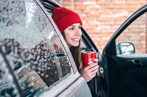 La giovane donna felice con una tazza termica rossa si siede in un'automobile nell'inverno