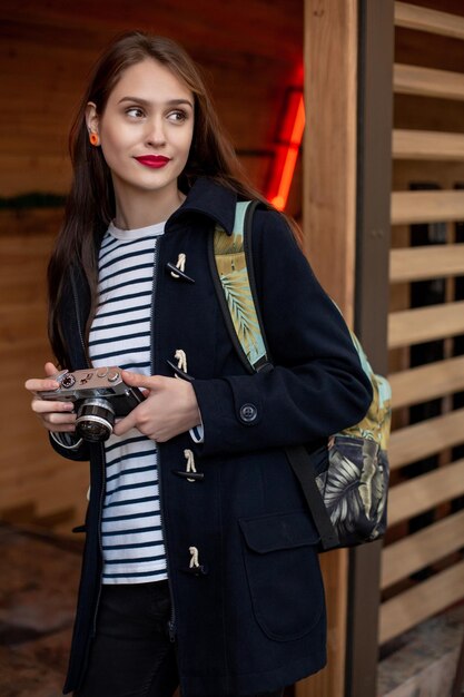 La giovane donna felice dei pantaloni a vita bassa tiene la macchina fotografica retrò. Divertirsi in città con la macchina fotografica, foto di viaggio del fotografo.