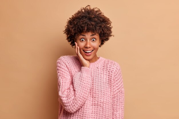 La giovane donna felice sorpresa con i capelli afro guarda con eccitazione e un ampio sorriso tiene la mano sul viso vestito con un maglione lavorato a maglia casual pone contro il muro beige dello studio