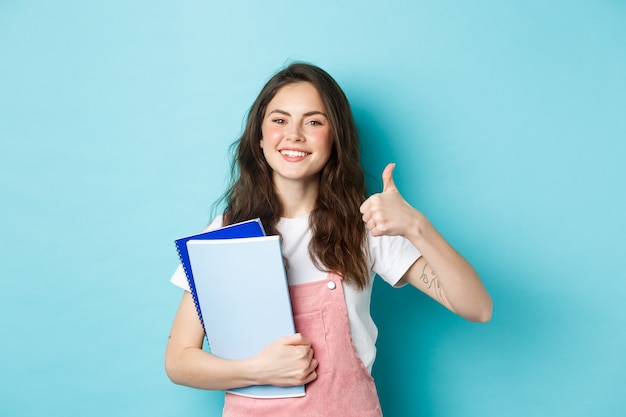 La giovane donna frequenta i corsi, la studentessa studia, tiene in mano i quaderni e mostra il pollice in su in segno di approvazione, raccomandando compagnia, in piedi su sfondo blu