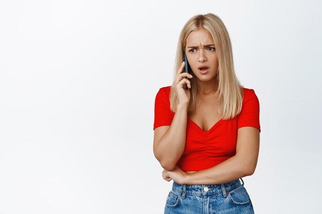La giovane donna frustrata riceve cattive notizie durante una telefonata che sembra sconvolta e scioccata mentre conversa sullo sfondo bianco dello smartphone