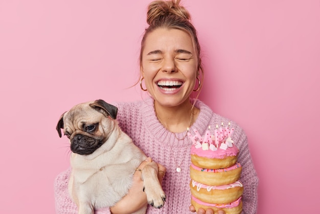 La giovane donna gioiosa ride felicemente tiene l'animale domestico preferito sulle mani deliziose ciambelle dolci con candele accese celebra il compleanno dei cani divertirsi insieme isolato su sfondo rosa studio