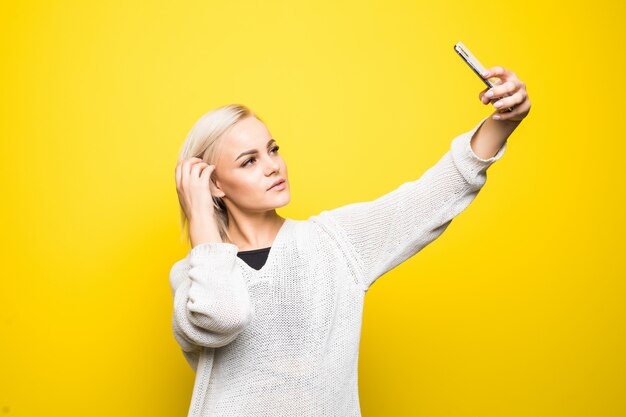 La giovane donna graziosa della signora in maglione bianco fa selfie sul suo smartphone su giallo