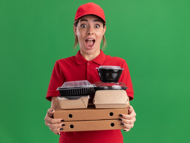 La giovane donna graziosa di consegna emozionante in uniforme tiene i pacchetti e i contenitori di cibo di carta sulle scatole della pizza isolate sulla parete verde