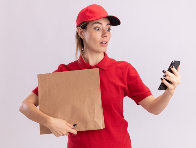 La giovane donna graziosa di consegna impressionata in uniforme tiene il pacchetto di carta e guarda il telefono isolato sulla parete bianca