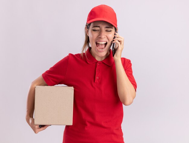 La giovane donna graziosa di consegna infastidita in uniforme tiene il cardbox e urla a qualcuno sul telefono isolato sulla parete bianca
