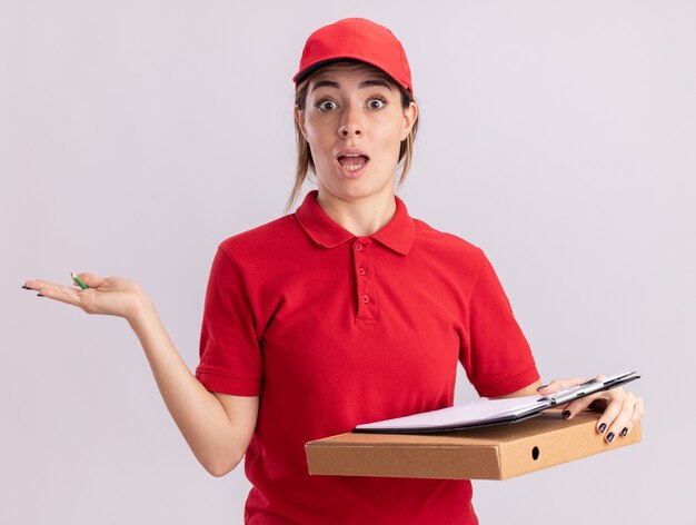 La giovane donna graziosa di consegna sorpresa in uniforme tiene la mano aperta e tiene la lavagna per appunti sulla scatola della pizza isolata sulla parete bianca