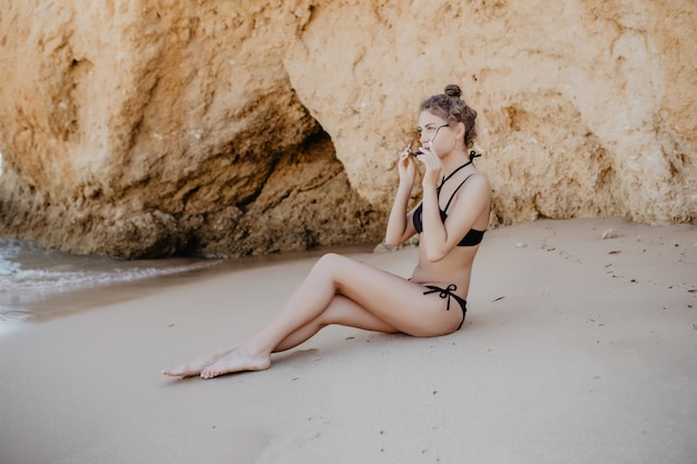 La giovane donna graziosa in bikini gode del sole sulla spiaggia rocciosa dell'oceano