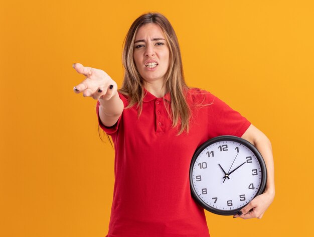 La giovane donna graziosa infastidita tiene l'orologio e indica nella parte anteriore con la mano isolata sulla parete arancione