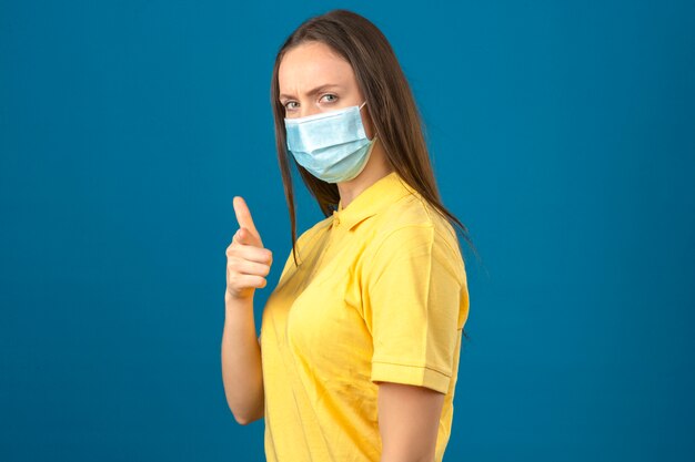 La giovane donna in camicia di polo gialla e maschera protettiva medica che indica il dito la macchina fotografica con il fronte serio che sta sul blu ha isolato il fondo