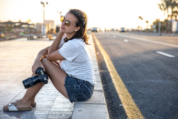 La giovane donna in estate in pantaloncini corti e una maglietta si siede sul marciapiede con una macchina fotografica al tramonto