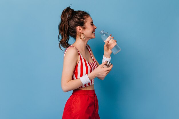 La giovane donna in vestito a strisce rosse ride e beve l'acqua dalla bottiglia sulla parete isolata