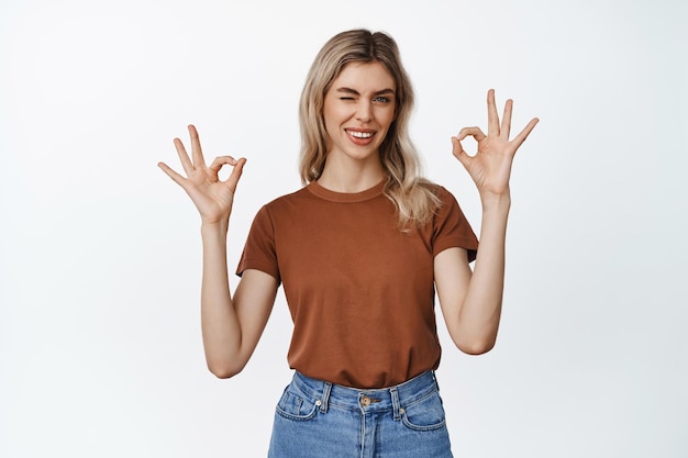 La giovane donna mostra il gesto di conferma ammiccando e facendo segni di approvazione assicurandoti che ti piace e approva il prodotto in piedi su sfondo bianco