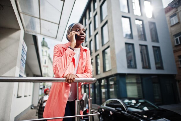 La giovane donna musulmana africana alta e snella attraente moderna alla moda in hijab o sciarpa di turbante parla sul telefono cellulare contro la città