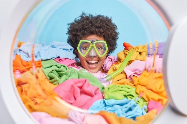 La giovane donna occupata dai capelli ricci allegra indossa la maschera per lo snorkeling per le immersioni ha molto bucato da fare fa i lavori domestici annegati in abiti multicolori contro il muro blu