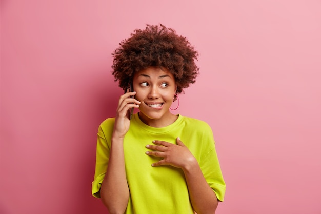 La giovane donna positiva con i capelli ricci parla tramite il telefono cellulare morde le labbra guarda da parte si sente felice vestita con una maglietta casual isolata sopra il muro rosa aspetta fino a quando l'amico non risponderà