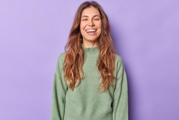 La giovane donna positiva con lunghi capelli ondulati vestita con un maglione casual si sente molto felice di essere di buon umore ascolta i complimenti isolati su sfondo viola e guarda qualcosa di divertente. Concetto di emozioni