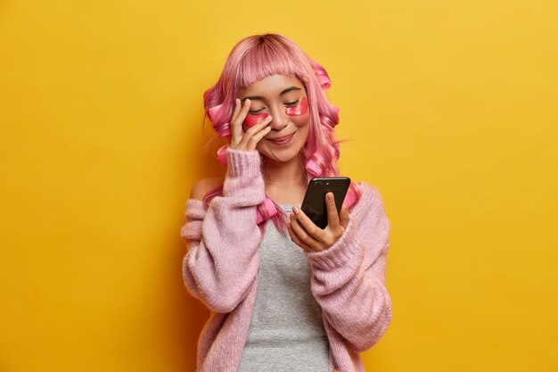 La giovane donna positiva ridacchia positivamente e guarda il display dello smartphone, legge notizie divertenti, ha lunghi capelli rosa, fa acconciatura, si preoccupa per la pelle