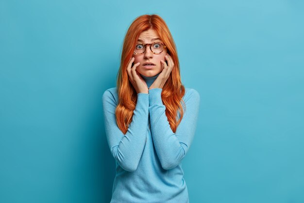 La giovane donna rossa scioccata preoccupata tiene le mani sulle guance e guarda sorprendentemente eccitata da notizie inaspettate vestite con abiti casual.