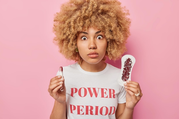 La giovane donna scioccata con i capelli ricci tiene il tampone quotidiano e il tampone usa un prodotto per l'igiene delle mestruazioni vestito con una maglietta isolata su sfondo rosa Donne giorni critici intimi e concetto di ciclo