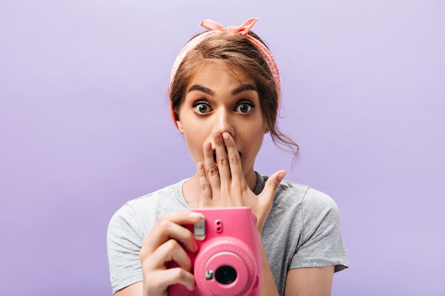 La giovane donna scioccata tiene la macchina fotografica rosa. Ragazza fresca con fascia alla moda estiva e acconciatura alla moda su sfondo isolato.