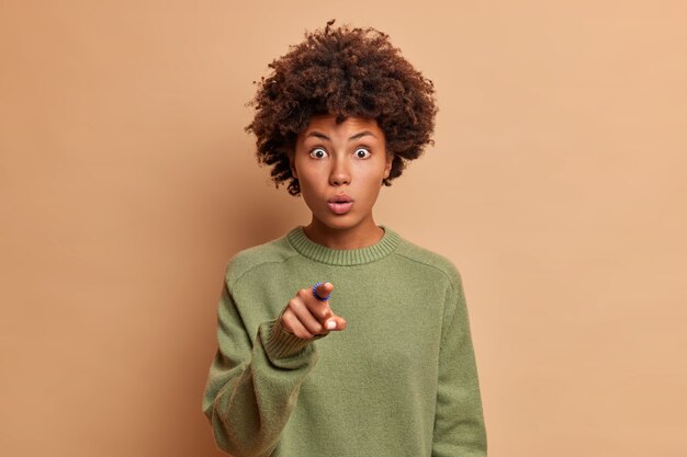 La giovane donna senza parole con i capelli ricci punta direttamente sul davanti trattiene il respiro vestito casualmente vede qualcosa di incredibile o mozzafiato isolato sul muro beige