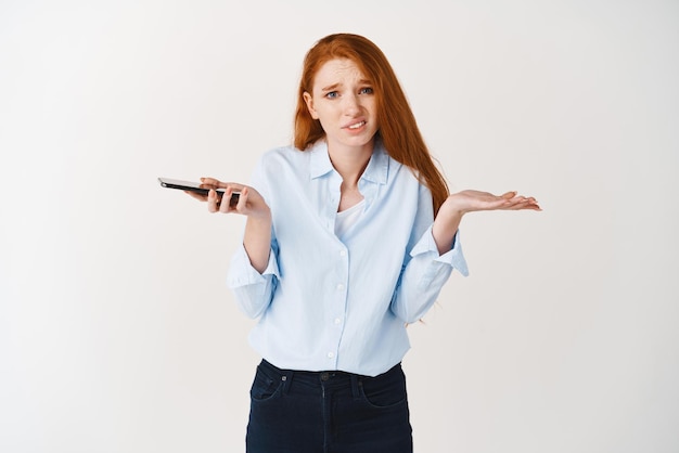 La giovane donna senza tracce con i capelli rossi e la camicia blu che scrolla le spalle la fotocamera che tiene lo smartphone e che sembra confusa non conosce lo sfondo bianco