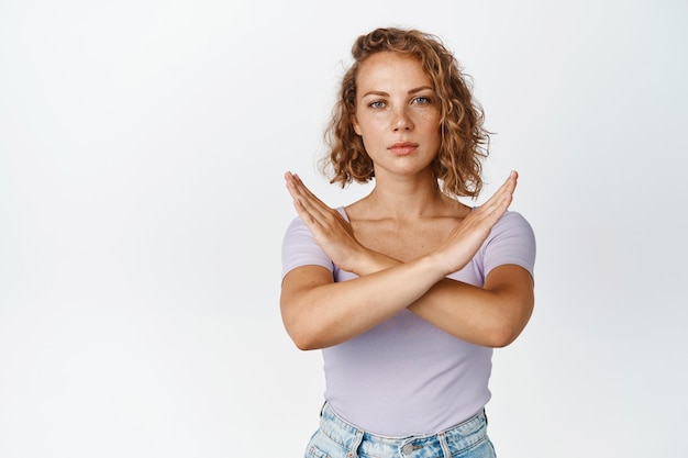 La giovane donna seria mostra il gesto di arresto incrociato, guardando perplesso, dicendo no, proibisce qualcosa, in piedi sul bianco.