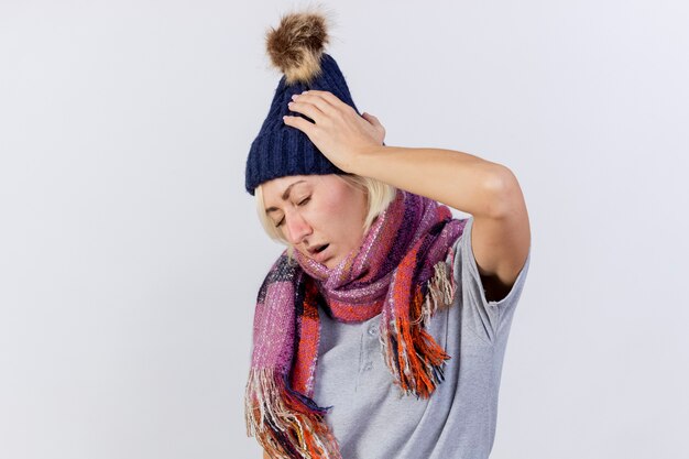 La giovane donna slava malata bionda dolorante che indossa il cappello e la sciarpa di inverno mette la mano sulla testa isolata sulla parete bianca con lo spazio della copia