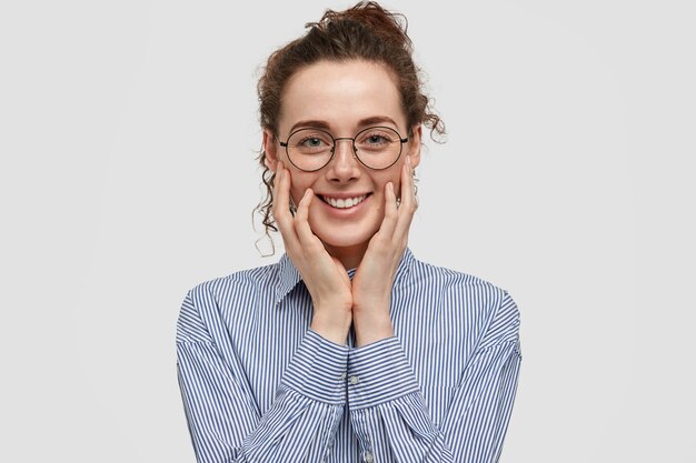 La giovane donna soddisfatta positiva ha un sorriso gentile, tocca le guance con le mani