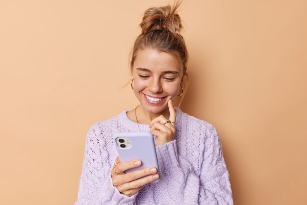 La giovane donna soddisfatta sorride felicemente mantiene il dito indice sulla guancia concentrato nel dispositivo smartphone legge un messaggio piacevole indossa un maglione casual isolato su sfondo beige. Tecnologia cellulare