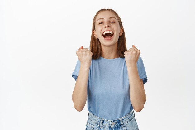 La giovane donna sollevata e felice urla dal successo e dal trionfo, fa pompare il pugno, gridando gioiosa, esulta come vincente, raggiunge l'obiettivo, in piedi contro il muro bianco