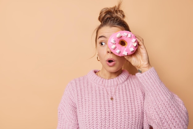 La giovane donna sorpresa guarda con un'espressione scioccata lontana tiene la bocca aperta copre gli occhi con una ciambella smaltata che va a mangiare uno spuntino delizioso indossa un maglione lavorato a maglia isolato su sfondo beige.