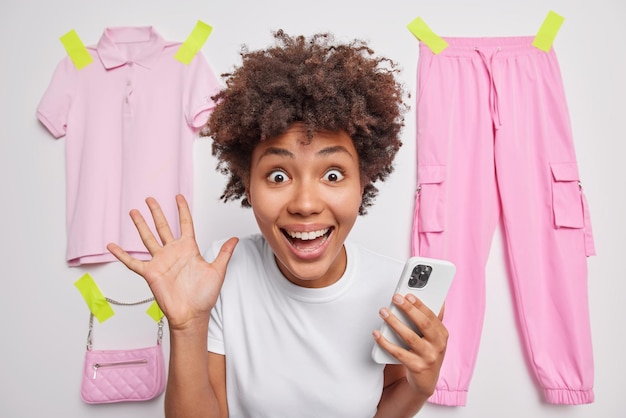 La giovane donna sorpresa positiva con i capelli ricci tiene il palmo alzato guarda con gioia la fotocamera utilizza lo smartphone per scorrere le pose di internet su uno sfondo bianco gli indumenti intonacati dietro