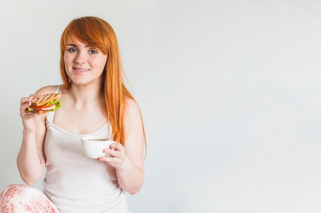 La giovane donna sorridente che tiene il panino e la tazza di caffè arrostiti contro fondo bianco