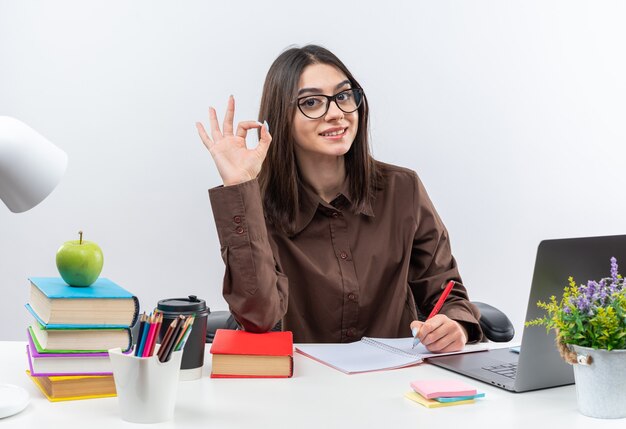 La giovane donna sorridente della scuola con gli occhiali si siede al tavolo con gli strumenti della scuola che mostrano un gesto ok