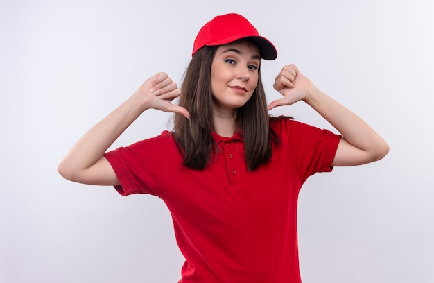 La giovane donna sorridente di consegna che porta la maglietta rossa in berretto rosso indica se stesso sulla parete bianca isolata