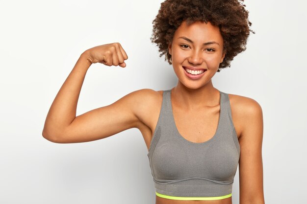 La giovane donna sorridente positiva della razza mista solleva il braccio muscoloso, mostra i bicipiti, ha un aspetto potente, sorride felice, vestita con reggiseno sportivo, isolato su sfondo bianco, dice guarda quanto sono forte.