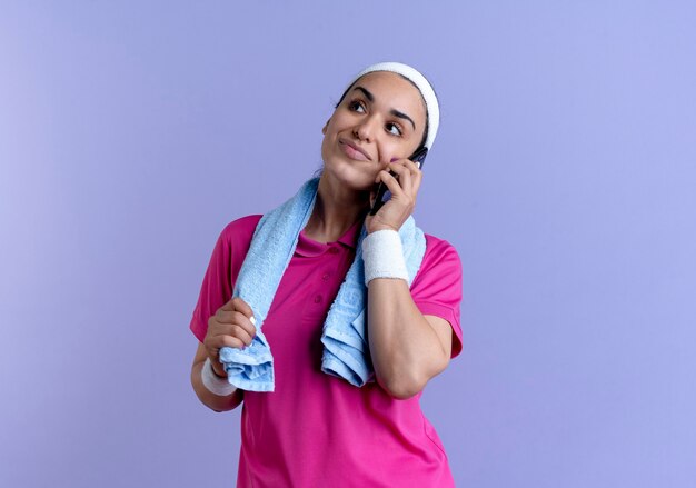 La giovane donna sportiva caucasica felice che porta la fascia ed i braccialetti tiene l'asciugamano intorno al collo che parla sul telefono sulla porpora con lo spazio della copia