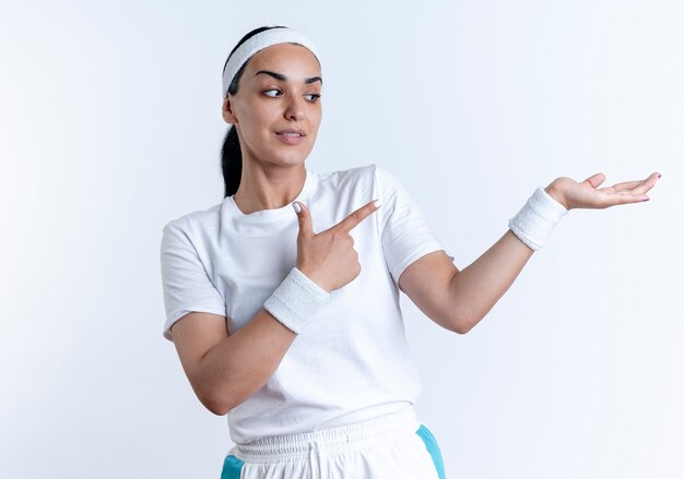 La giovane donna sportiva caucasica fiduciosa che indossa la fascia e i braccialetti indica la mano vuota isolata su spazio bianco con lo spazio della copia