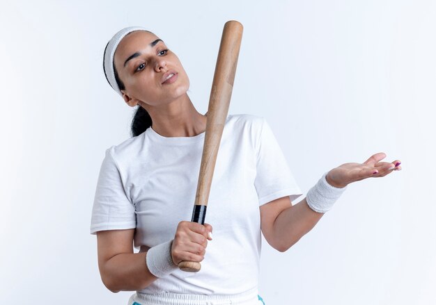 La giovane donna sportiva caucasica fiduciosa che indossa la fascia ed i braccialetti tiene il pipistrello che guarda e che indica al lato isolato su spazio bianco con lo spazio della copia