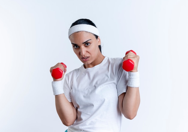 La giovane donna sportiva caucasica furiosa che indossa la fascia ed i braccialetti tiene i manubri isolati su spazio bianco con lo spazio della copia