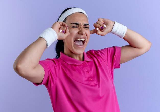 La giovane donna sportiva caucasica infastidita che indossa la fascia ed i braccialetti chiude le orecchie che esaminano il lato isolato sullo spazio viola con lo spazio della copia