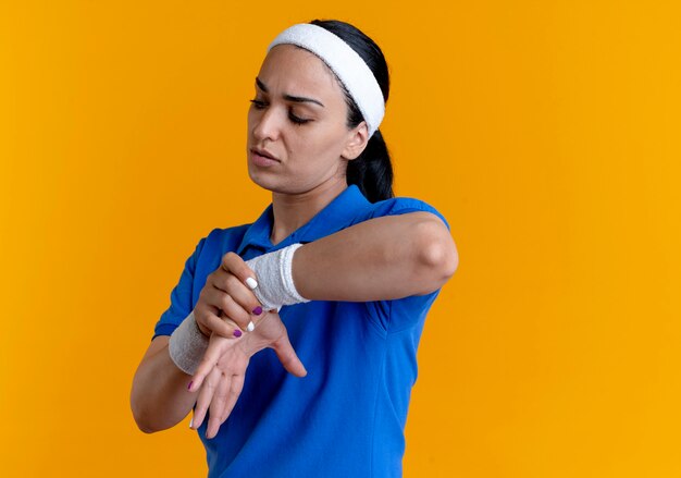 La giovane donna sportiva caucasica infastidita che indossa la fascia ed i braccialetti tiene la mano sull'arancio con lo spazio della copia