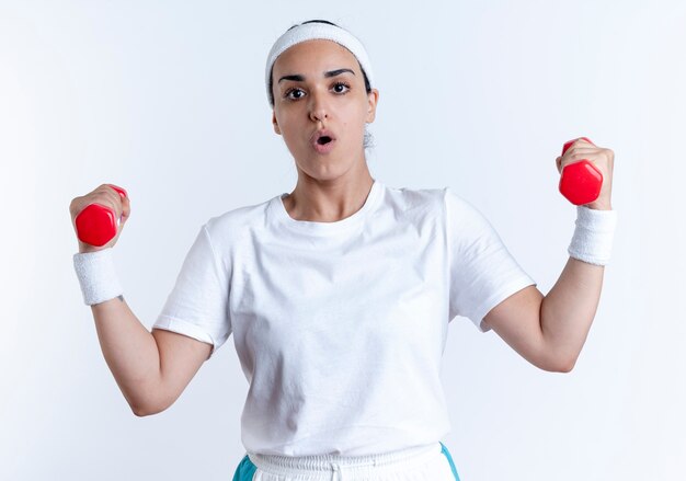 La giovane donna sportiva caucasica sorpresa che indossa la fascia ed i braccialetti tiene i dumbbells isolati su spazio bianco con lo spazio della copia