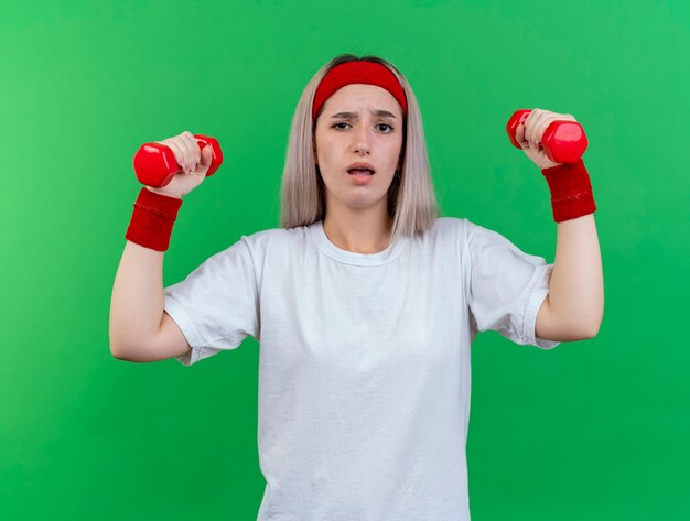 La giovane donna sportiva senza tracce con le parentesi graffe che indossa la fascia e i braccialetti tiene i manubri isolati sulla parete verde